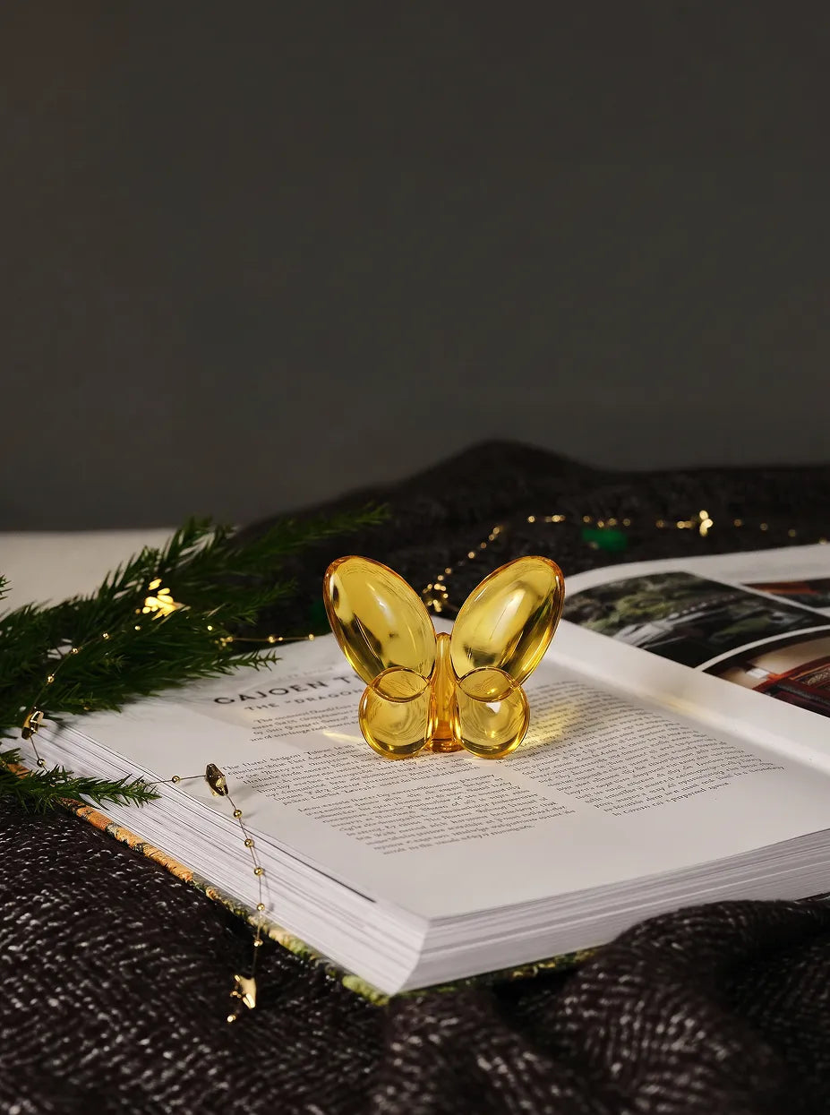 Official lifestyle image of the RESONG Butterfly Ornament - Amber, an elegant amber glass decorative sculpture displayed on an open book in a contemporary home décor setting.