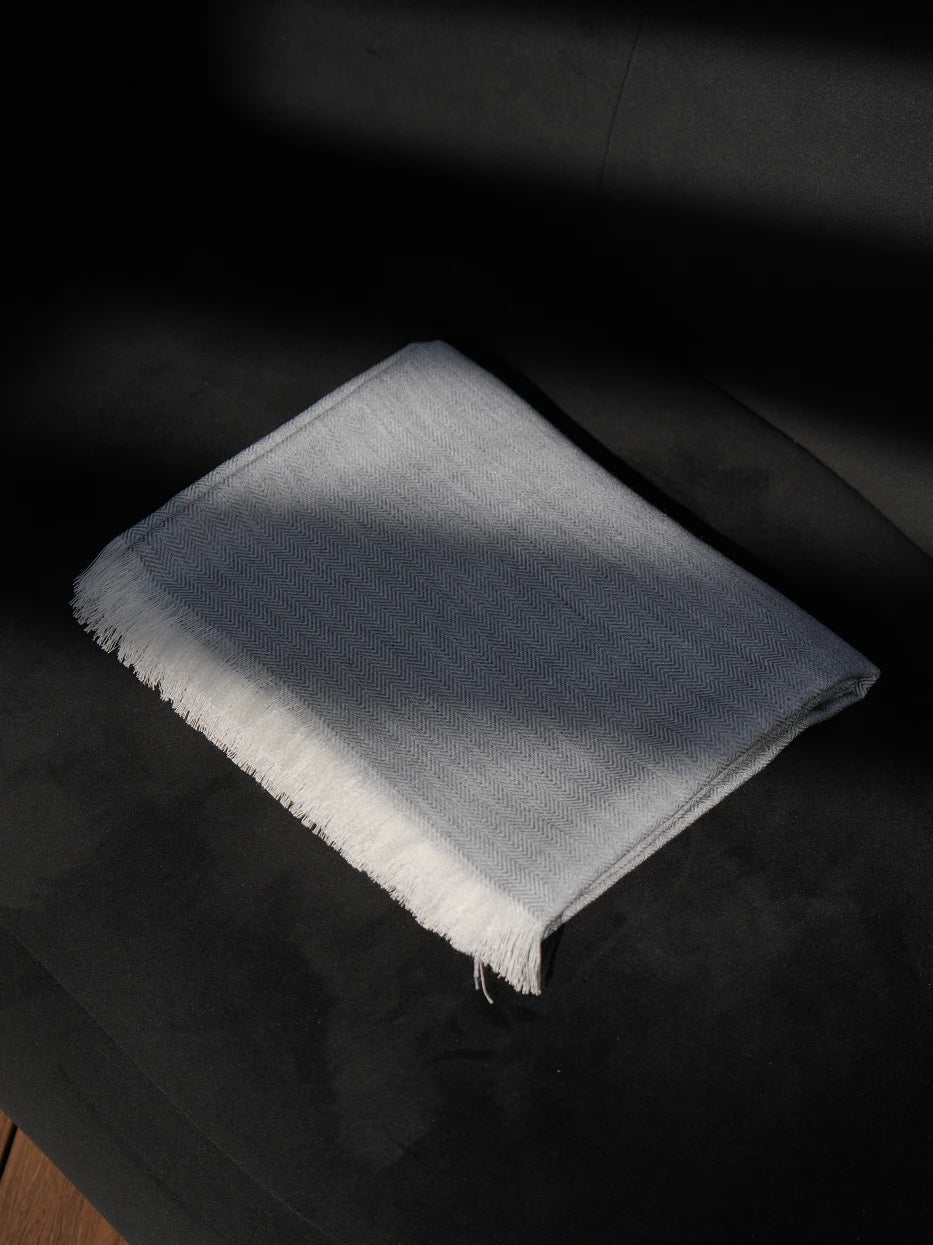 Minimalist interior still shot of folded CHENZHI herringbone wool throw placed on curved black accent armchair on solid wood flooring, cool high-contrast match between soft gray textile and dark furniture, lightweight silky breathable blend fabric without bulky texture, great decor for entryway lounge, reading nook, hotel rest corner, modern dark tone interior matching