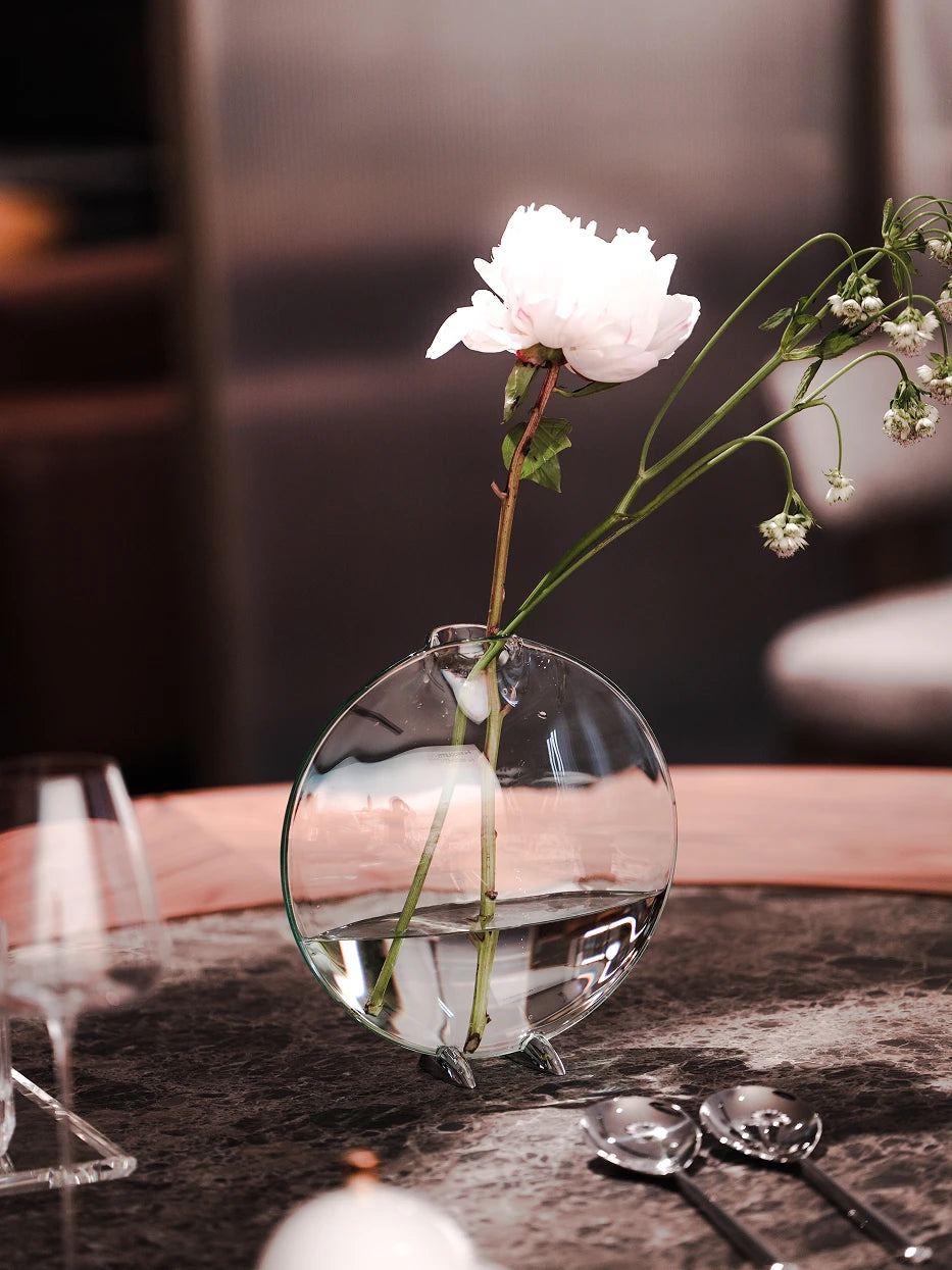 Close-up texture still life shot of medium Fishmouth vase with peony on dark marble coffee table, soft side light highlights transparent glass & alloy feet craftsmanship