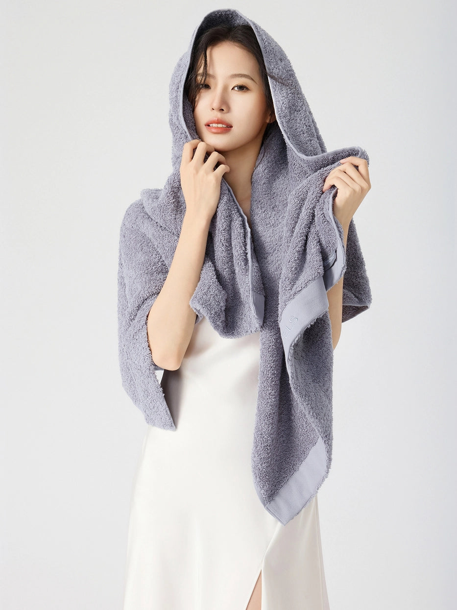 Woman wearing RESONG gray cotton terry bath towel draped over head like a hood, one hand gently touching chin showing fluffy absorbent texture, white slip dress, white studio background