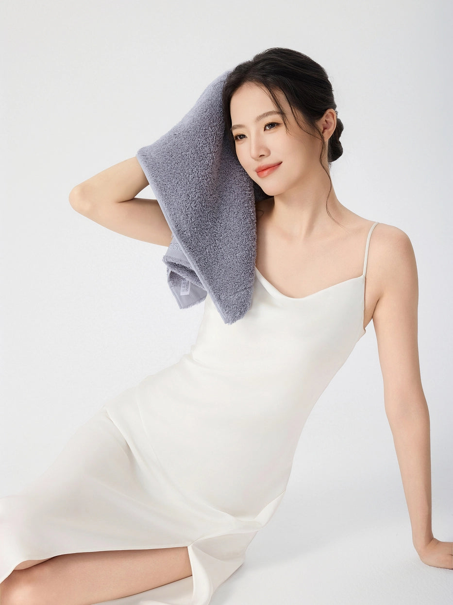 Woman sitting on floor in white slip dress, gently pressing RESONG gray cotton terry face towel against her cheek, soft fluffy face towel gift set, white studio background