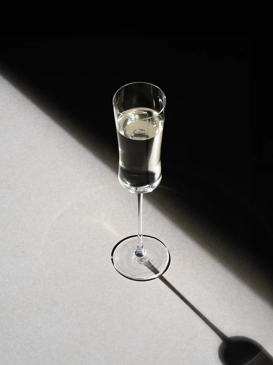 Top-down minimalist close-up of RESONG HOME 1833 curved crystal champagne flute filled with white sparkling wine, long soft shadow on matte light background