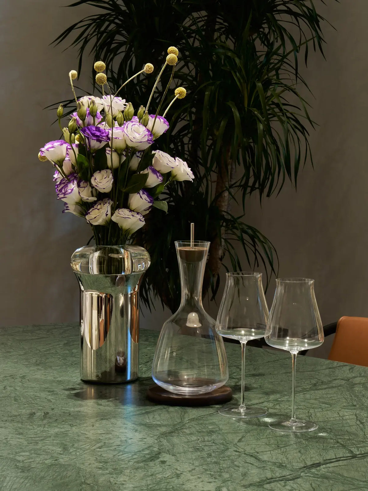 Elegant home dining lifestyle photo of empty RESONG HOME 1833 wine decanter matched with pair of 1833 red wine glasses, styled with metal flower vase and green plants on emerald marble table