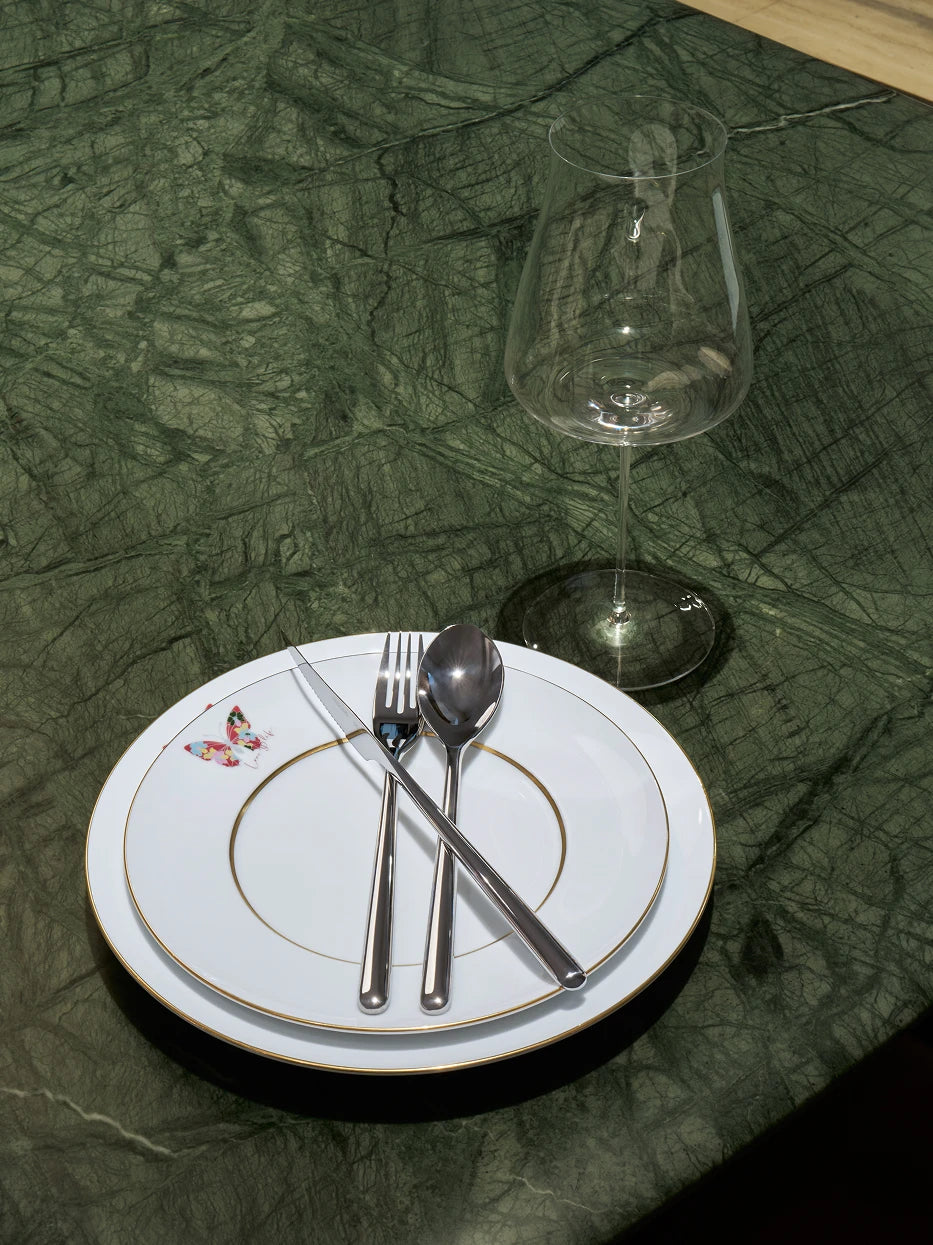 Minimal home table setting on dark green marble surface, stacked butterfly porcelain plates with stainless steel cutlery and wide-belly wine goblet