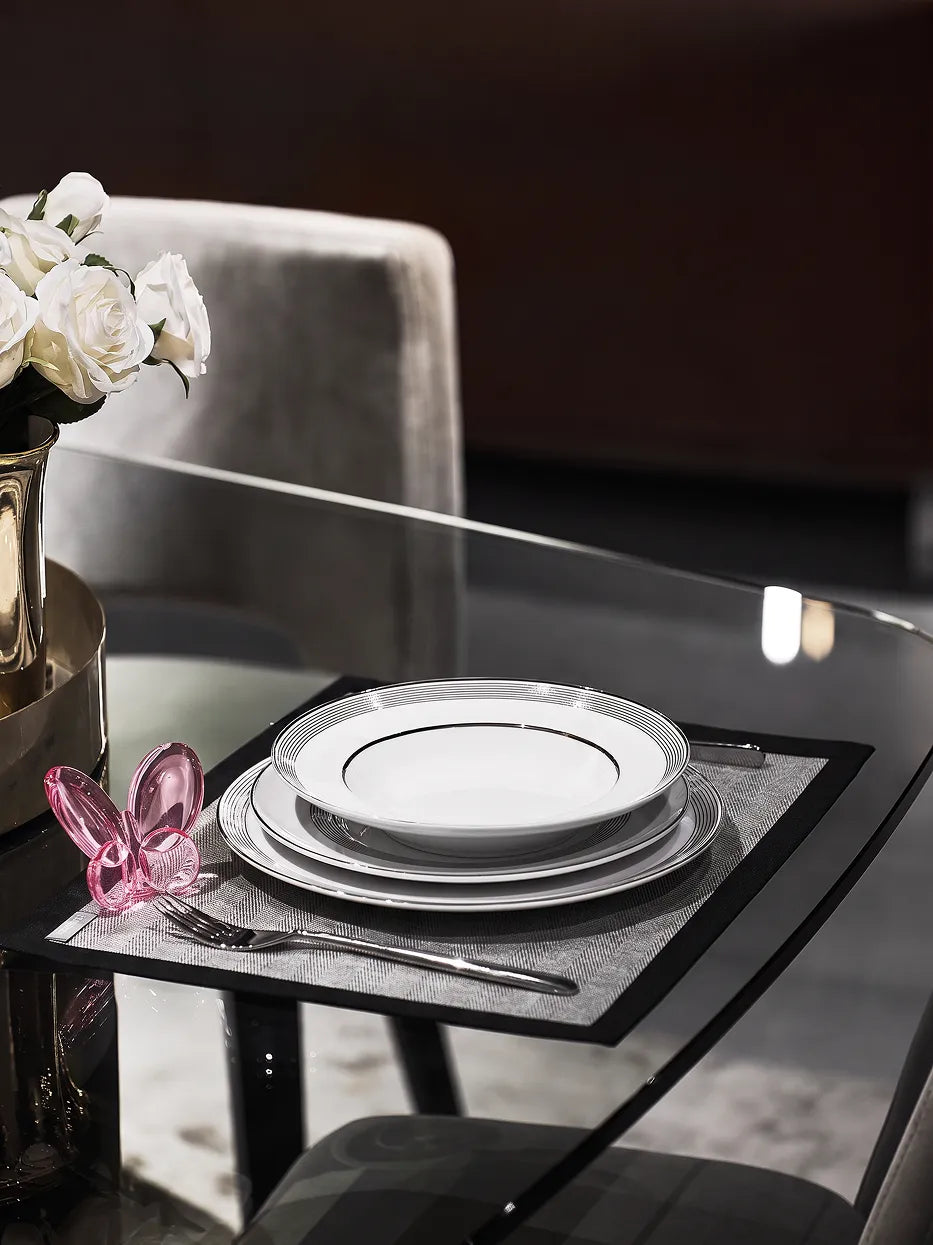 Light luxury home dining layout shot of stacked ERYA black-silver deep & flat plate set, matched with rose gold glass butterfly ornament, white rose vase and full metal cutlery on transparent glass tabletop