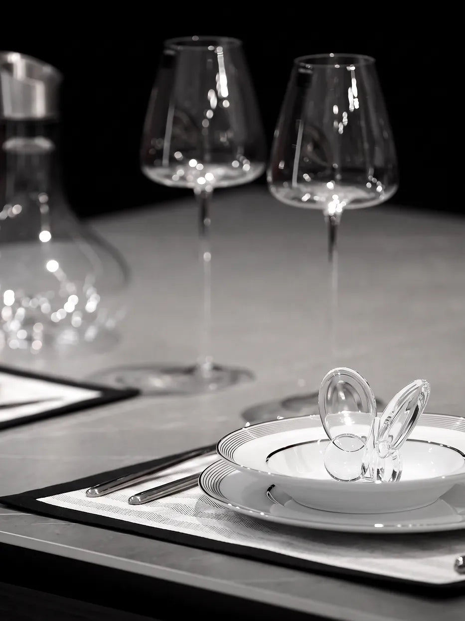 Monochrome dark studio close-up still life of ERYA black silver plate set with tall wine goblets and clear glass butterfly ornament, soft reflective light highlighting porcelain glaze texture