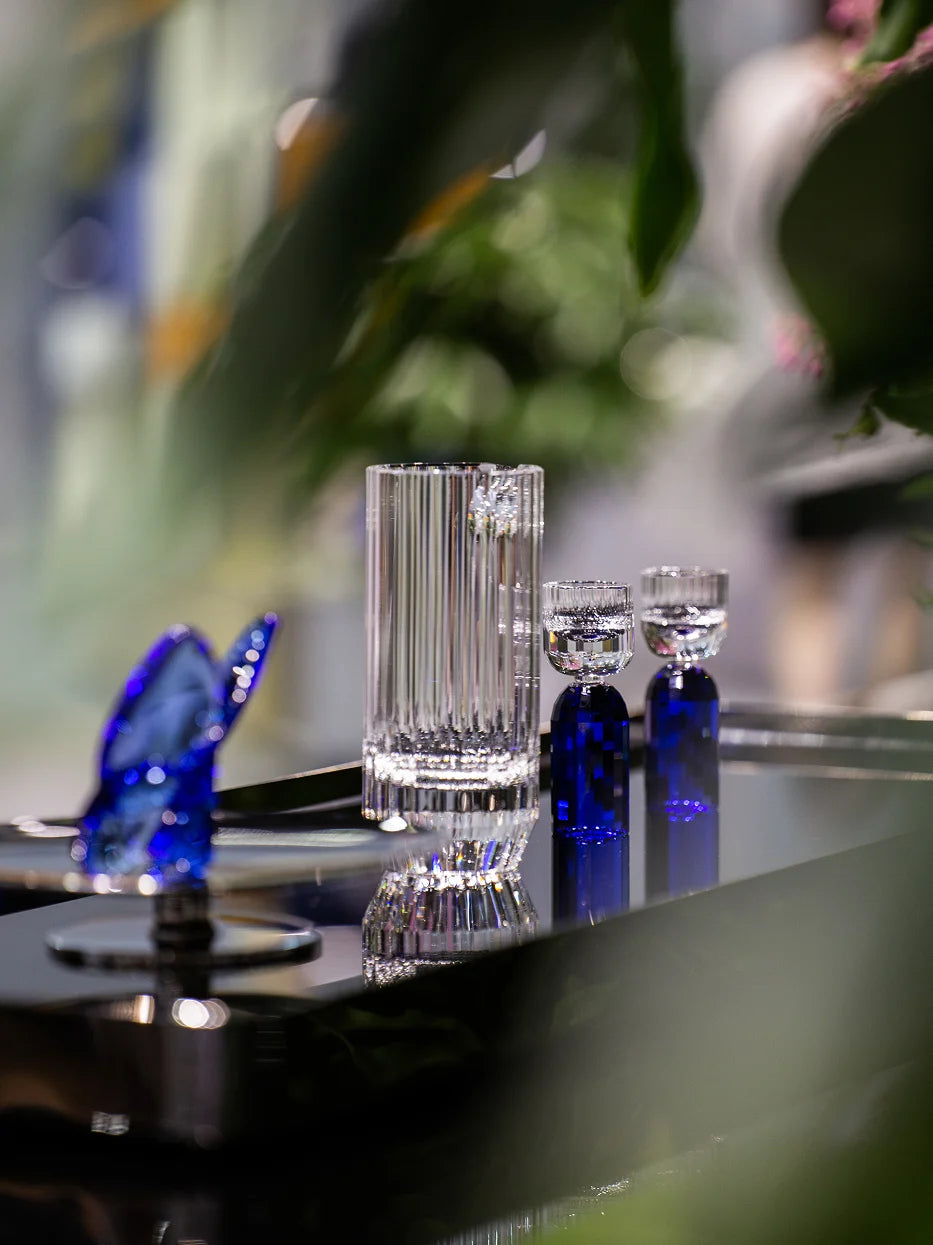 Dark luxury lifestyle shot of HAOYING sapphire blue baijiu crystal set on reflective metal tray, blurred green foliage and bokeh light background