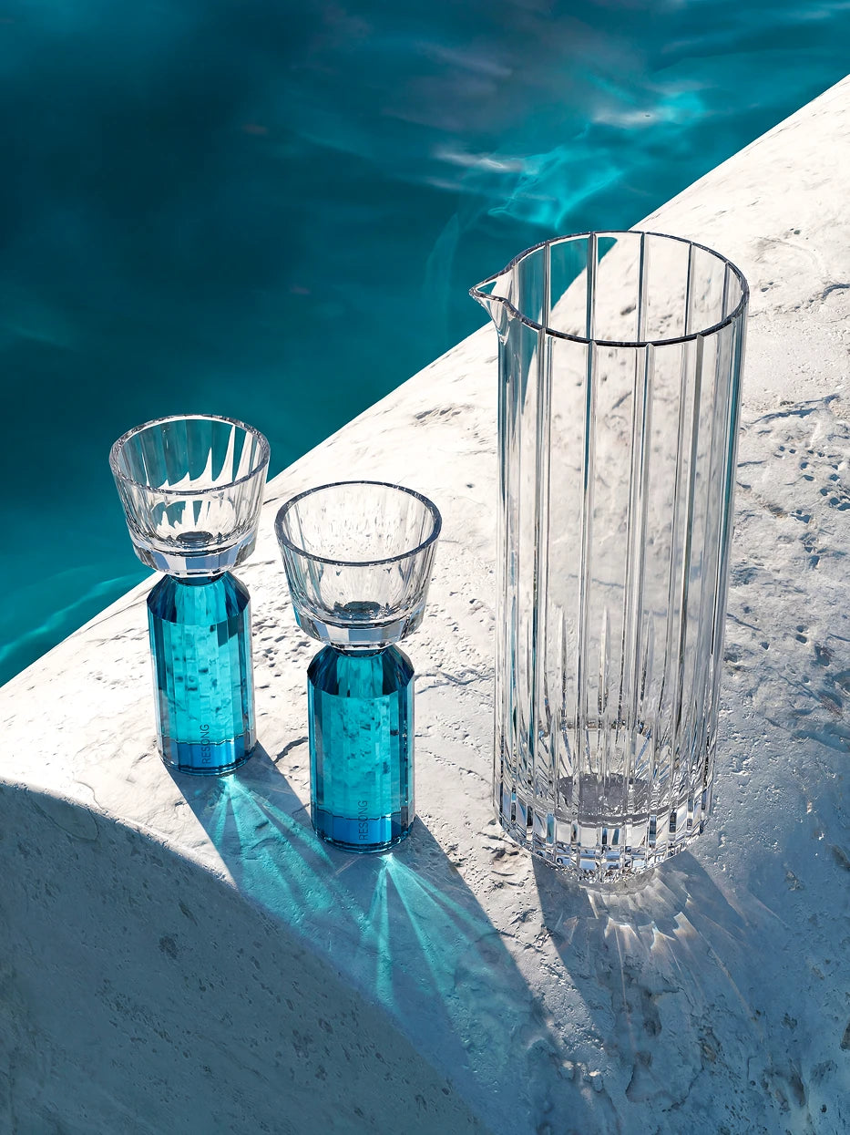 Vacation-style elegant lifestyle photo of HUAZUN lake blue crystal baijiu set on light stone poolside terrace, clear blue pool water background, bright natural sunlight casting striped glass shadows