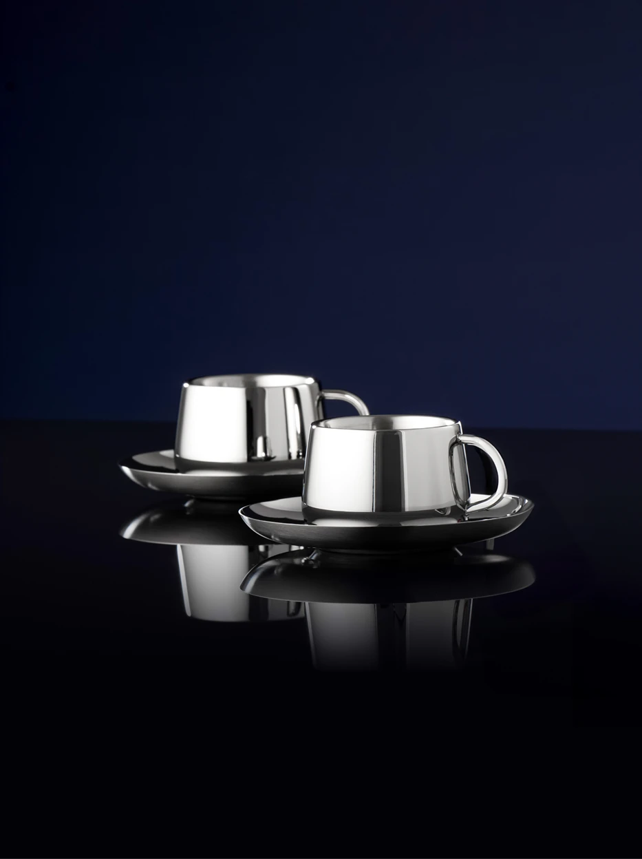 Studio still life of two sets of SHEDI small mirror steel cup & saucer, solid deep blue background highlighting clear reflective texture of polished stainless steel