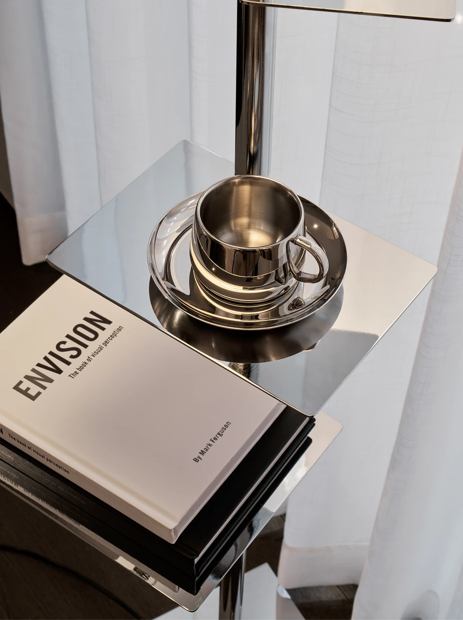 Light luxury side table shot of SHEDI mirror steel small cup & saucer placed on multi-layer metal stand, stacked art books and sheer white curtain as soft backdrop