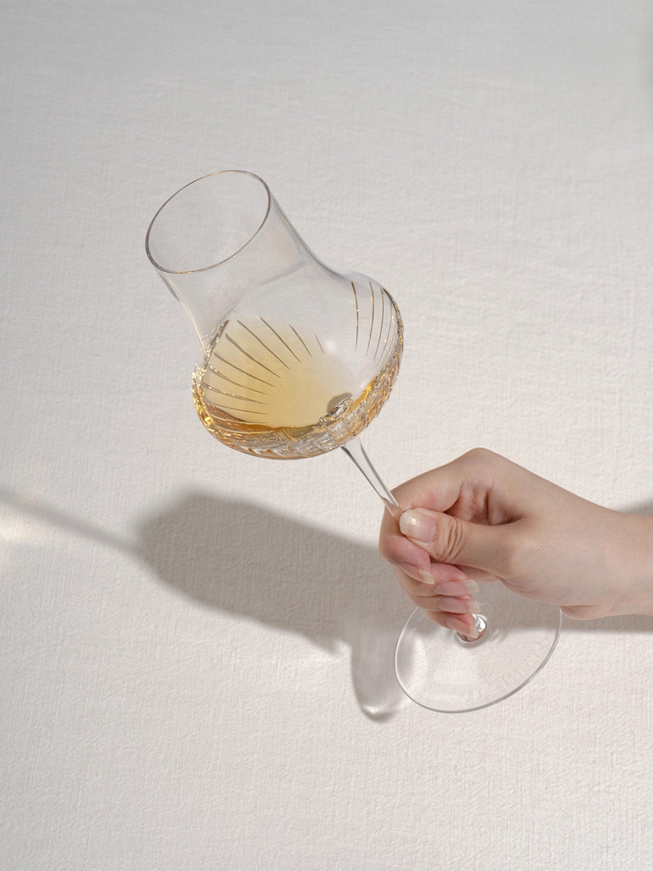 Soft minimalist lifestyle close-up of hand holding TRIUMPH carved crystal stem glass filled with pale white wine, matte textured light grey wall background