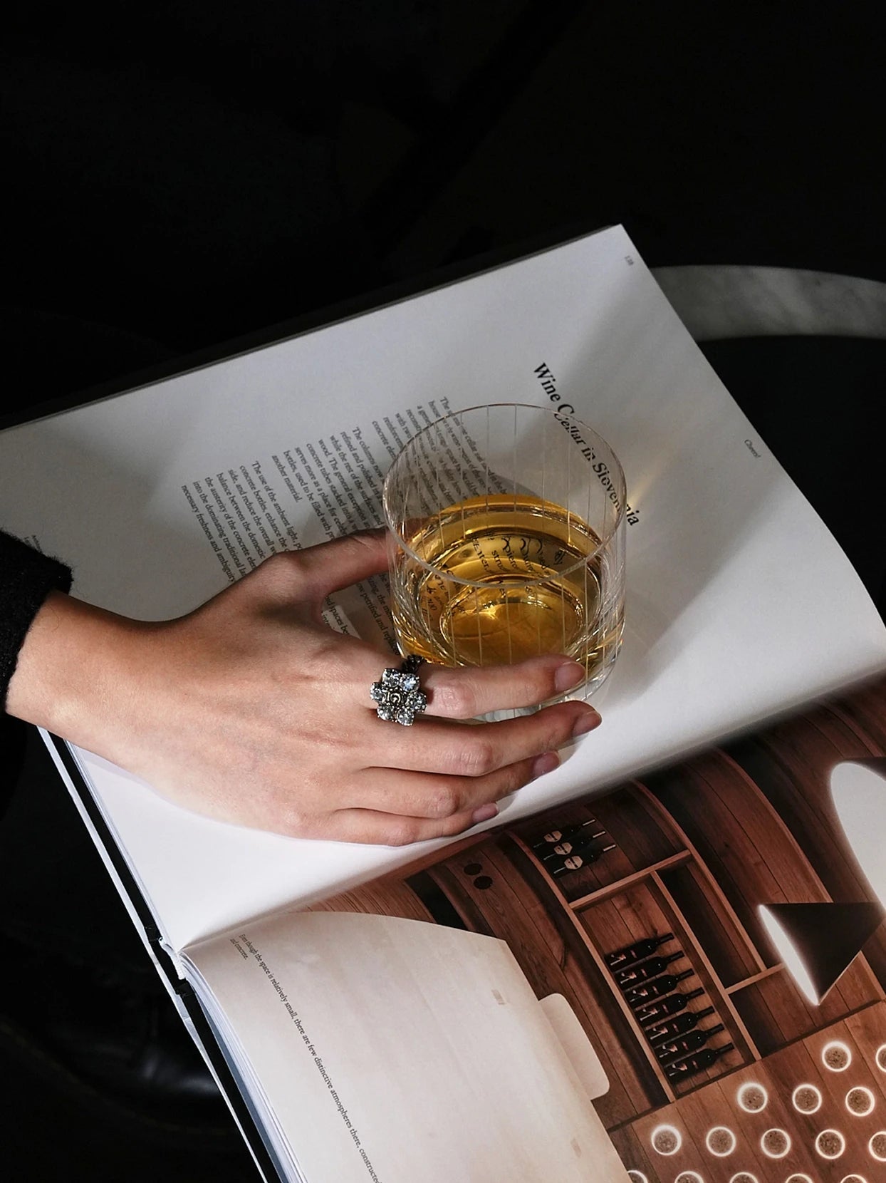 Luxury close-up shot of hand with diamond ring holding RESONG HOME XUYU vertical line crystal whiskey glass with amber liquor, resting on wine cellar art magazine page