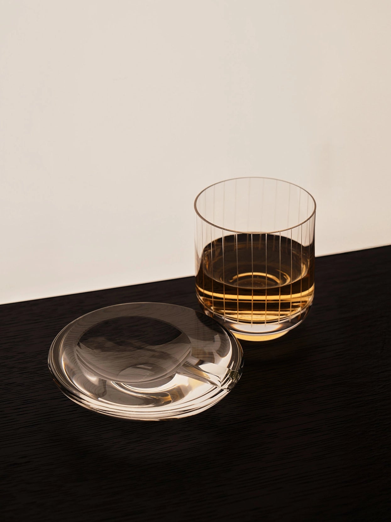 Minimal studio close-up of RESONG HOME XUYU etched whiskey tumbler with golden liquor, matched with original crystal saucer on matte dark wooden table, soft side shadow