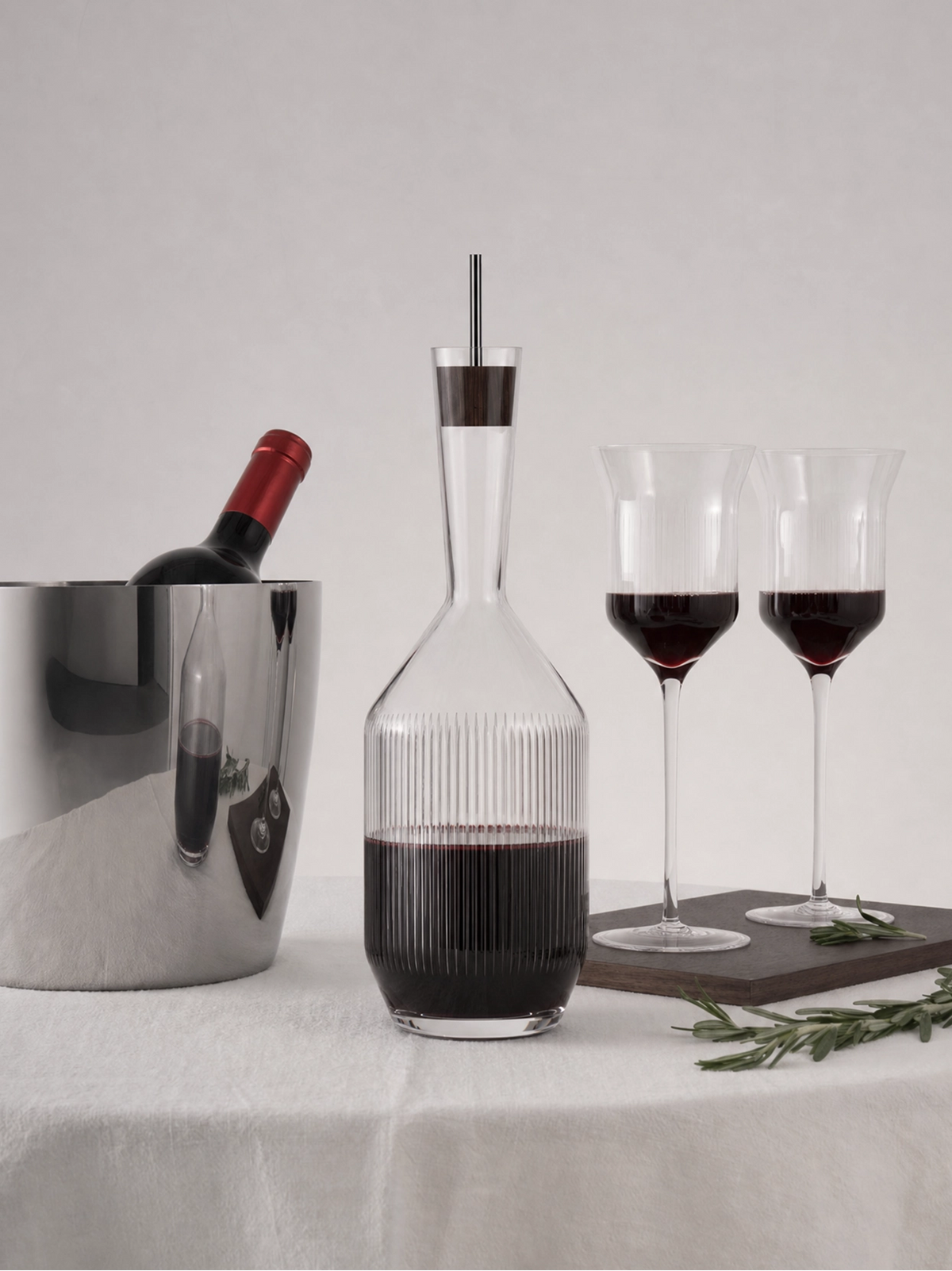 Soft dining lifestyle shot of RESONG HOME RXUYU ribbed crystal decanter filled with red wine, matched with vertical ribbed wine flutes, metal ice bucket & rosemary garnish on off-white linen tablecloth