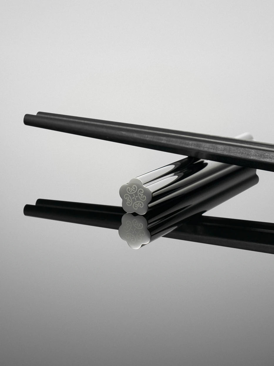 Dark tone close-up shot focusing on carved plum blossom scroll pattern of LINGLONG chopstick rest, black wood chopsticks laid on top to highlight mirror finish and delicate engraving