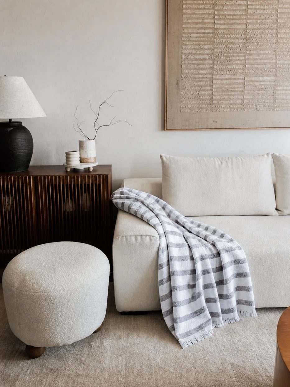 Wabi-sabi full living room furnishing layout shot, MUJIE gray striped linen fringe throw draped on beige fabric sofa, matched with solid wood slatted sideboard and round boucle ottoman, textured stripe blanket adds layered softness to plain sofa, perfect decor for villa living room, homestay lounge, large flat apartment, cream neutral tone interior styling