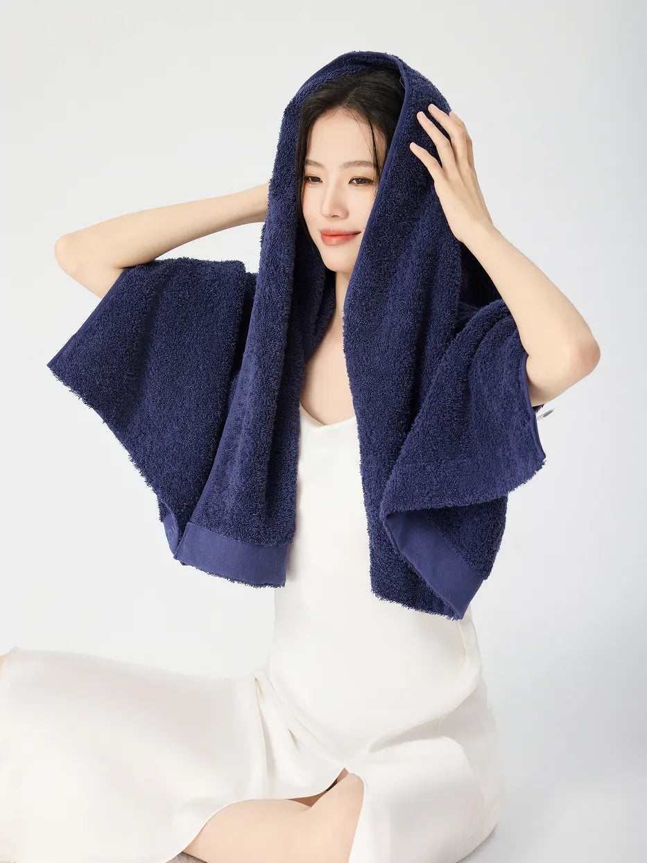 Woman holding RESONG navy blue cotton terry bath towel over head like a hood, wrapping wet hair after shower showing fluffy absorbent texture, white slip dress, white studio background