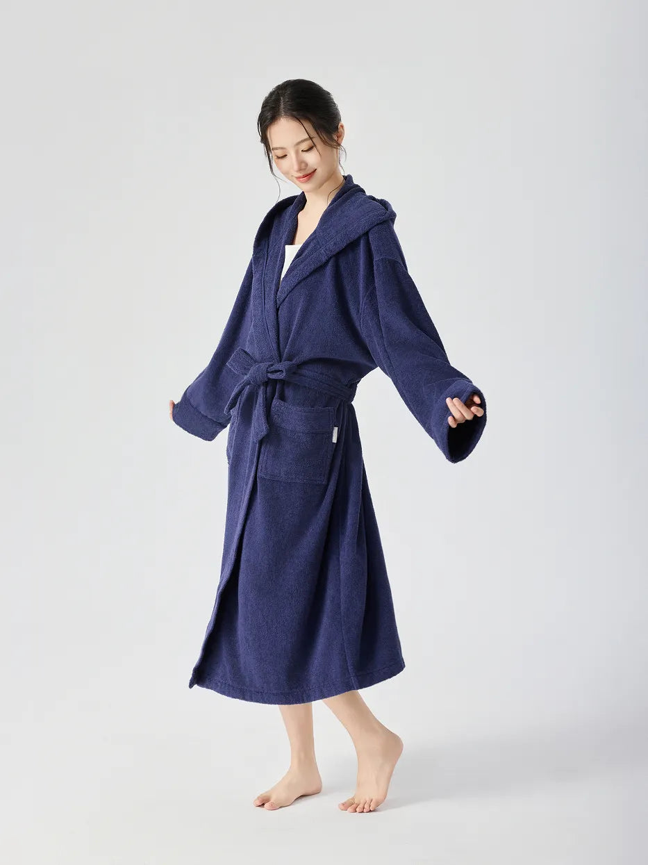Barefoot woman wearing RESONG navy blue hooded terry cloth bathrobe, full length, standing with arms slightly open showing relaxed fit, matching belt tie and patch pockets, soft absorbent cotton, white studio background