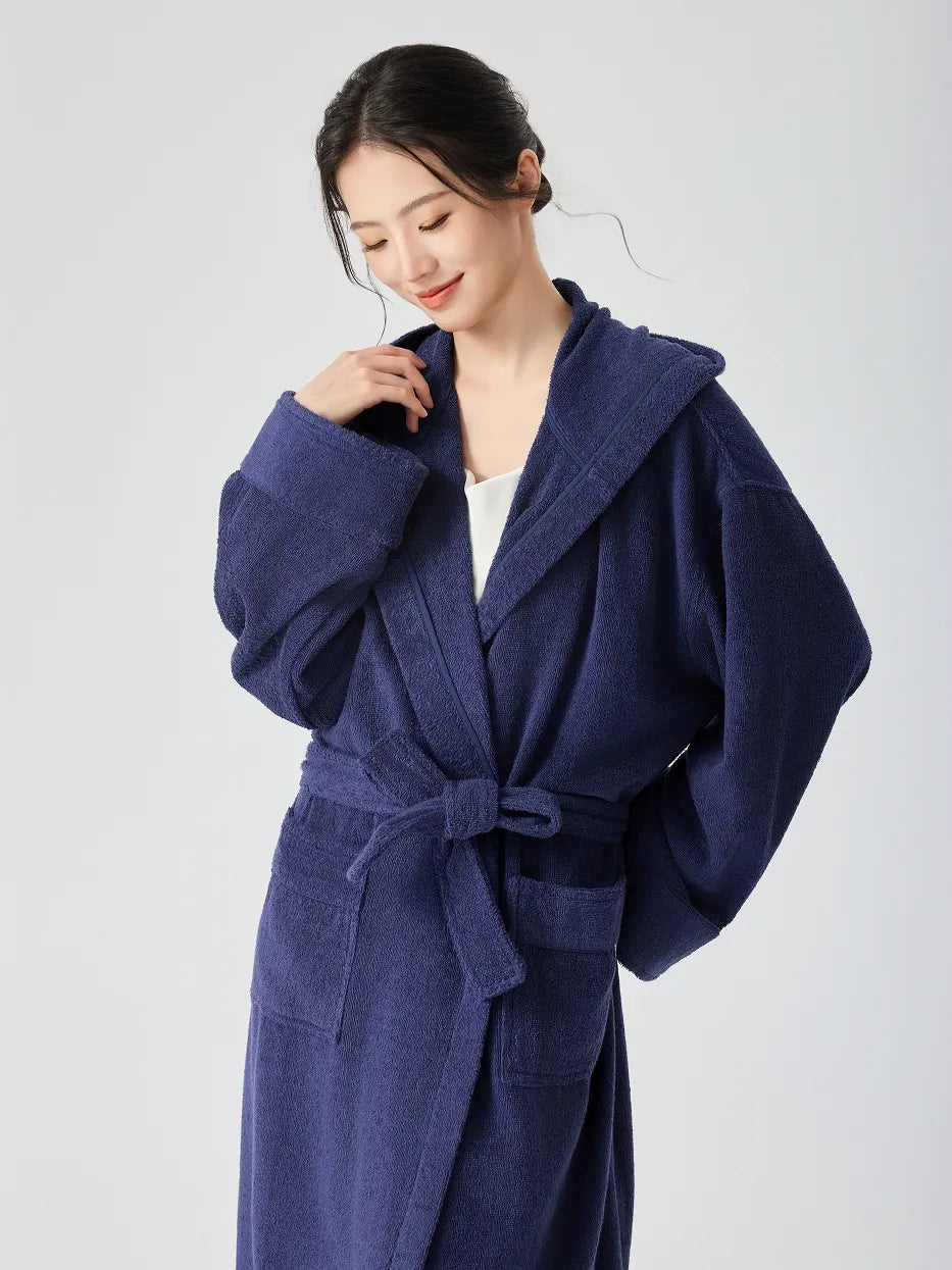 Woman wearing RESONG navy blue hooded terry cloth bathrobe, half body, smiling softly and looking down, one hand touching hood edge, matching belt tie, soft absorbent cotton, white studio background