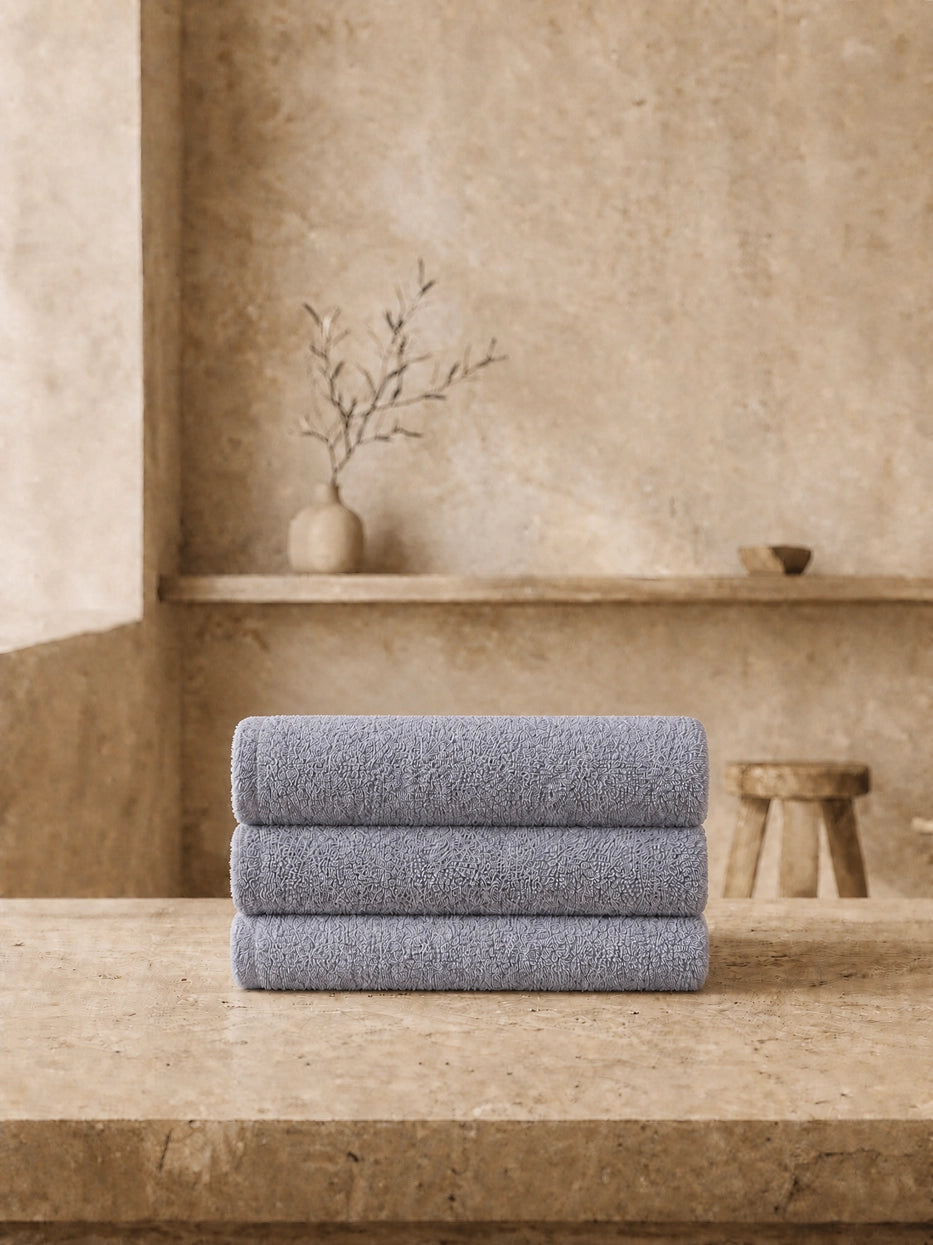 Wabi-sabi still life shot of three vertically stacked grey face towels on beige micro-cement countertop, unified solid grey tone soft and calm, matched with dried branch pottery decor to create minimalist bathroom vibe, shows full gift set display matching effect