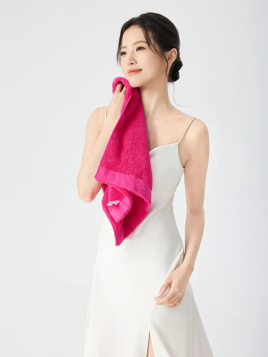 Woman in white slip dress gently pressing RESONG rose red cotton terry face towel against chin and neck, embroidered RESONG label on towel border visible, soft fluffy face towel on model, white studio background