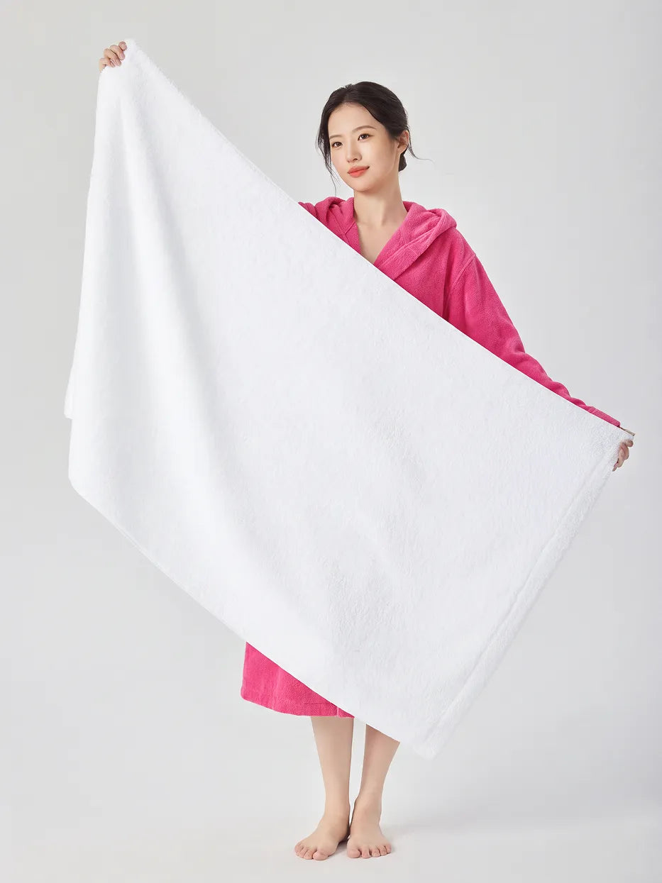 Barefoot woman wearing RESONG rose red hooded terry cloth bathrobe, holding a large white cotton bath towel fully open to show its size, luxury bath towel and bathrobe gift set, white studio background