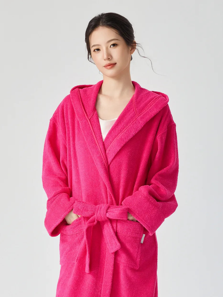 Woman wearing RESONG rose red hooded terry cloth bathrobe, half body front view, hands in patch pockets, matching belt tie, looking at camera, soft absorbent cotton, white studio background