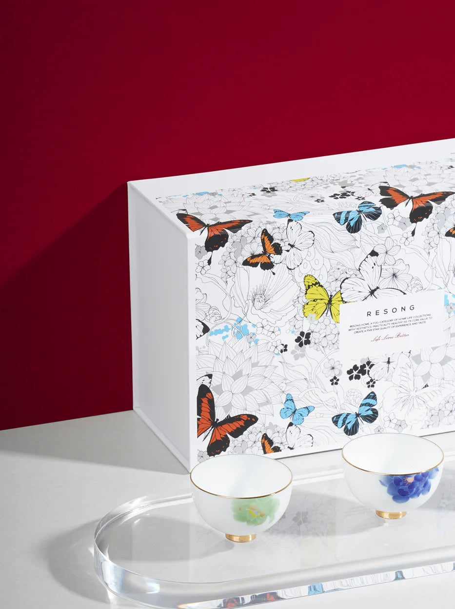 Display lifestyle shot of RESONG UNDERGLAZE MULTICOLORED ceramic tea cup set, two gold-rim floral teacups with transparent acrylic tray beside full-color butterfly printed gift box against deep red wall, premium tea gift for daily tea tasting & holiday gifting