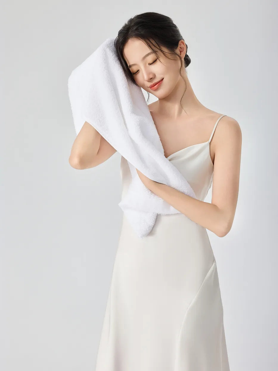 Woman in white slip dress, eyes closed gently pressing RESONG white cotton terry face towel against cheek to dry face, soft fluffy face towel on model, white studio background
