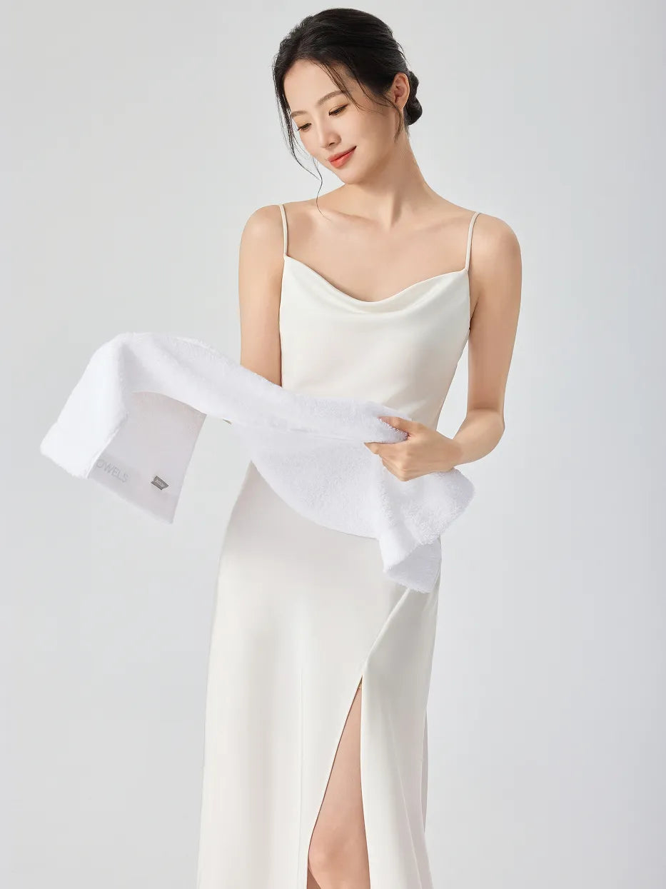 Woman in white slip dress looking down, unfolding RESONG white cotton terry face towel to show inner woven label, soft fluffy face towel on model, white studio background
