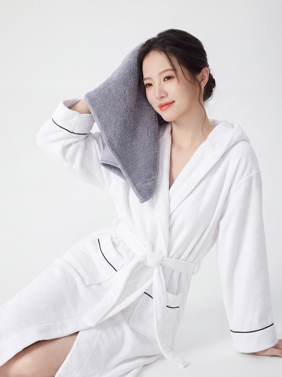 Woman sitting in RESONG white hooded terry cloth bathrobe with black piping, gently pressing a grey cotton bath towel against side of face, luxury bath towel and bathrobe gift set, white studio background