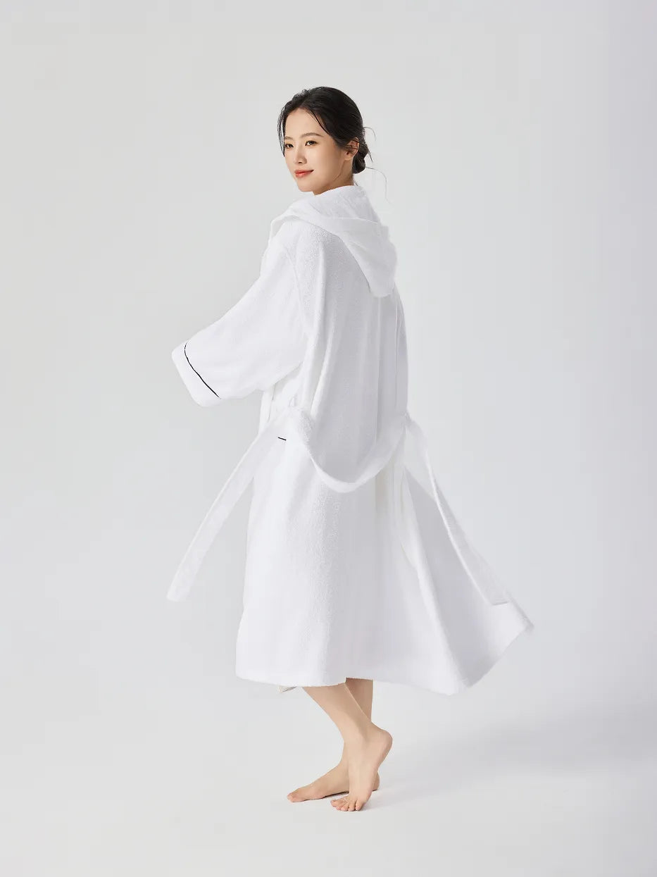 Barefoot woman turning back over shoulder wearing RESONG white hooded terry cloth bathrobe, hem flowing, showing hood back and navy piping on cuffs, relaxed long fit, white studio background