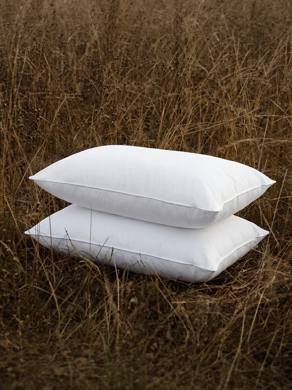 Two YUNGAN white goose down pillows stacked in wild dry grass, natural healing outdoor scene showing fluffy plump texture