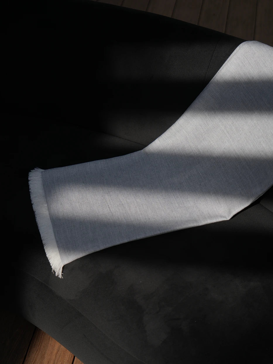 Interior drape texture shot of CHENZHI linen TENCEL wool blend gray throw blanket on black sofa, showing delicate herringbone woven texture and loose handmade white tassel fringe detail, visible silky luster of TENCEL™, breathable linen weave and soft wool mixed touch, neatly cut natural white fringed edges, displays premium multi-fabric craft and smooth drape tactile texture for home textile product display