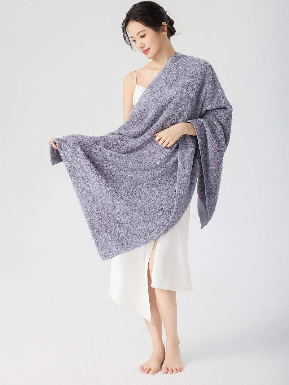 Barefoot woman standing draped in RESONG gray cotton terry bath towel over one shoulder, gently holding towel corner showing fluffy absorbent texture, white slip dress, white studio background
