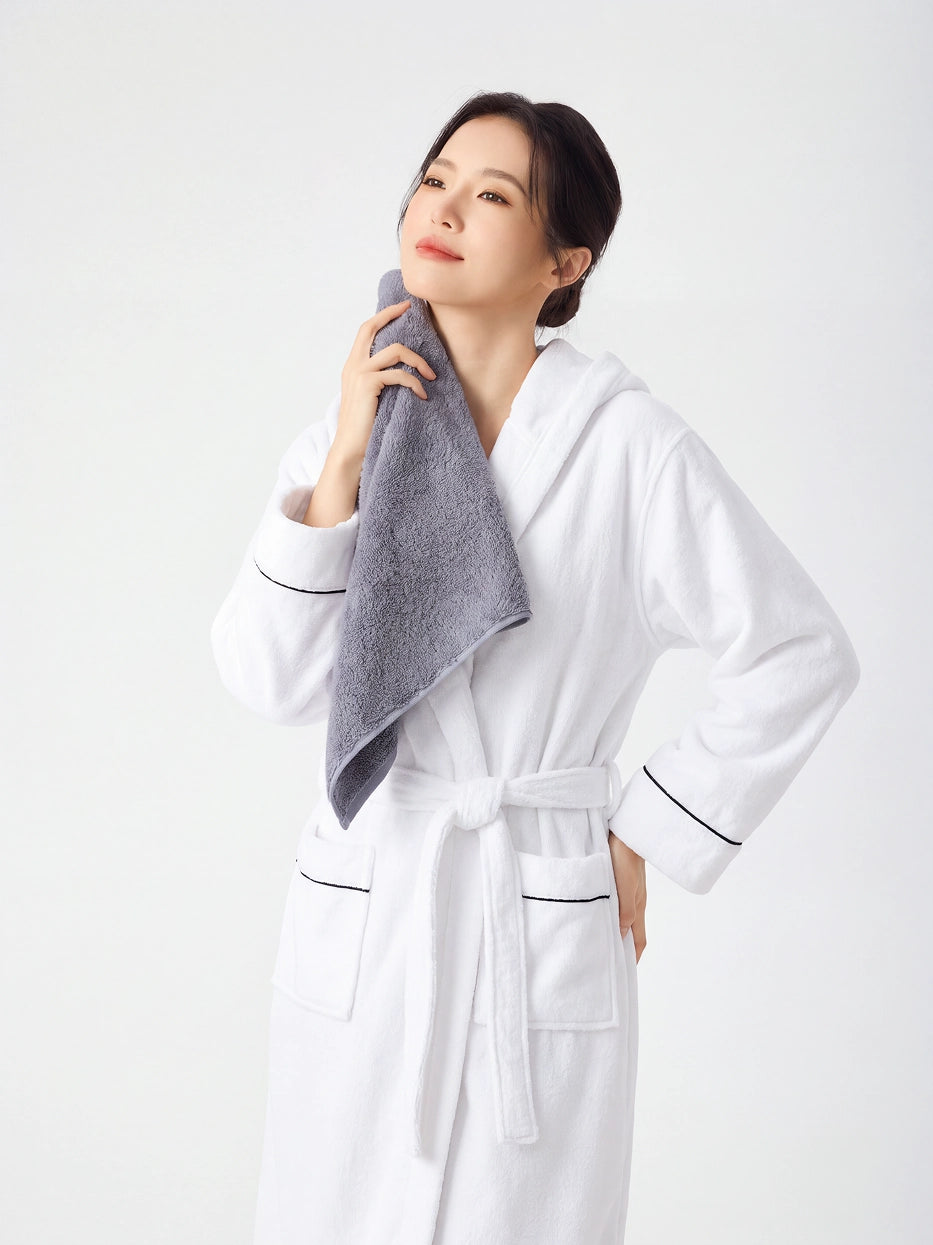 Woman in RESONG white hooded bathrobe with black piping, eyes closed pressing a gray cotton terry face towel under chin and against neck, soft fluffy face towel gift set, white studio background
