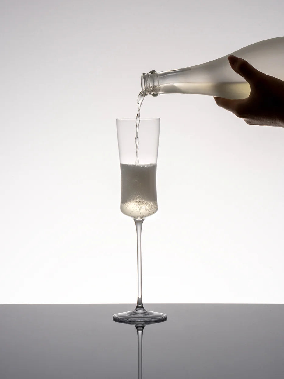 Minimal studio shot of RESONG HOME 1833 curved champagne flute being poured with sparkling wine, reflective dark mirror surface product photography