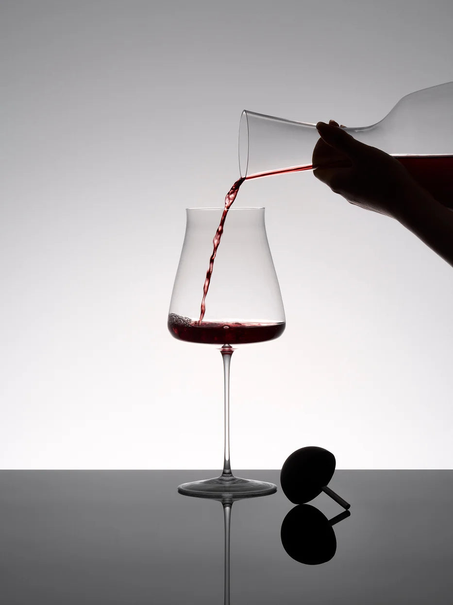 Studio product photo of RESONG HOME 1833 elegant large red wine goblet being poured red wine from crystal decanter, reflective black tabletop with wine stopper