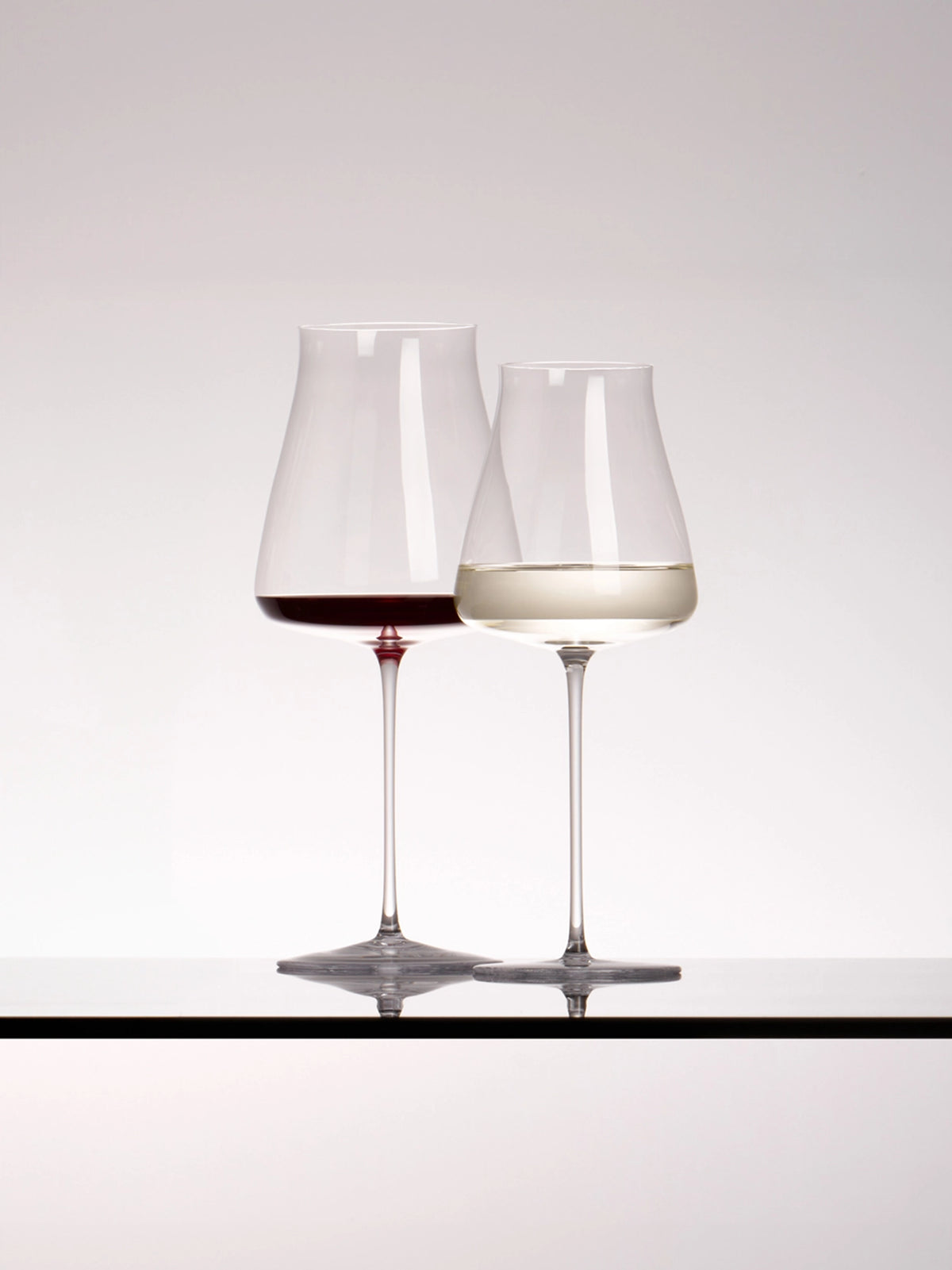 Clean studio display of two RESONG HOME 1833 slim crystal wine goblets, one filled with red wine, one with white wine, clear glass table reflective surface