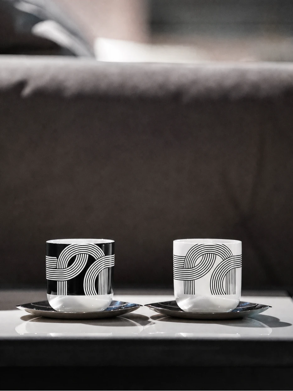 Minimalist modern living room shot of ALK black & white ceramic cup set on glossy reflective coffee table, dark grey leather sofa as soft blurred background