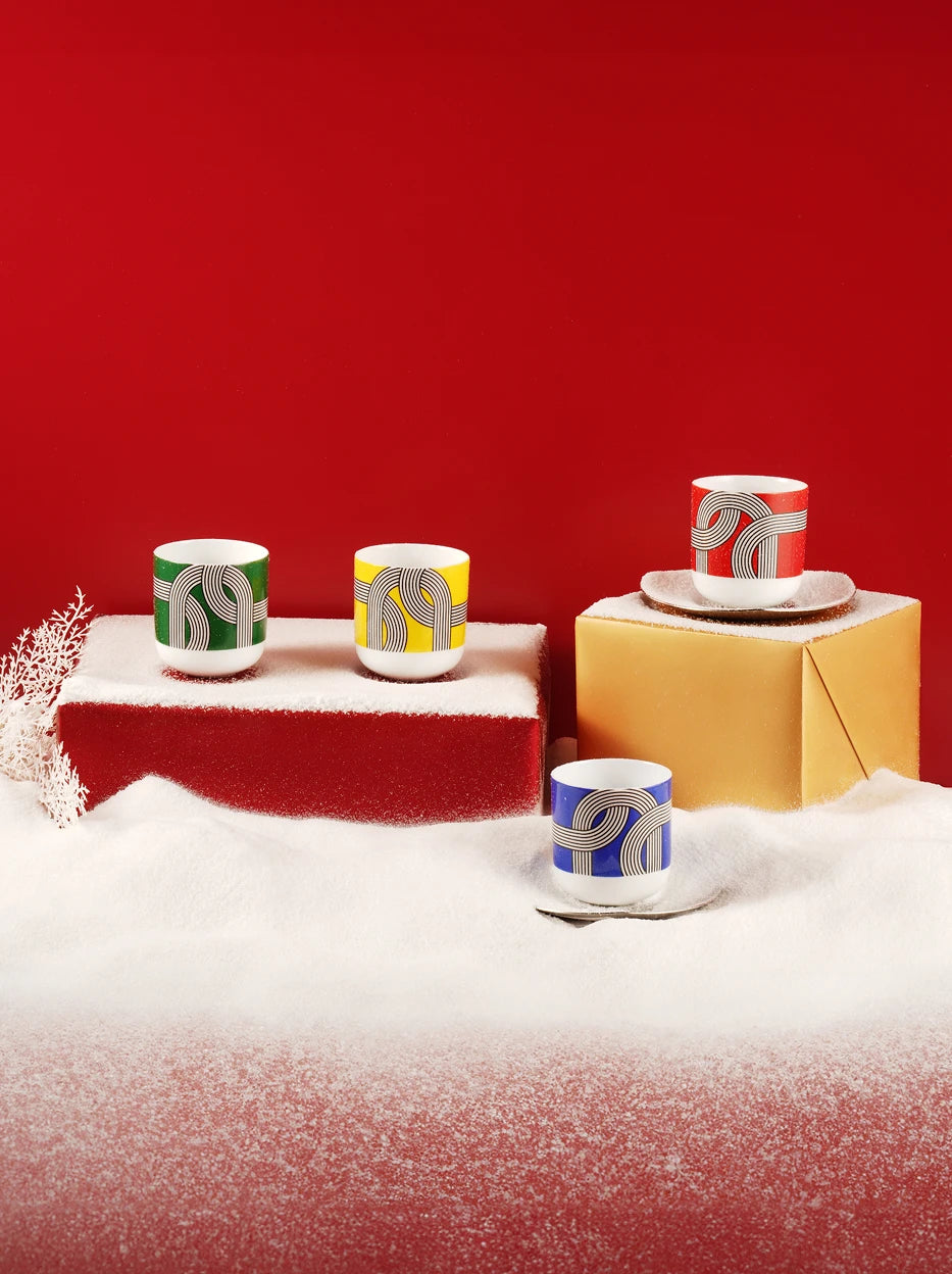 Festive winter lifestyle shot of ALK four-color ceramic cups arranged on red & gold gift boxes covered with artificial snow, solid deep red background for holiday gift display