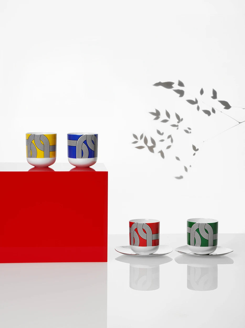 Minimal artistic studio display of four ALK geometric print ceramic cups, laid on glossy red cubic platform and reflective white table, leaf shadow decoration on blank white wall