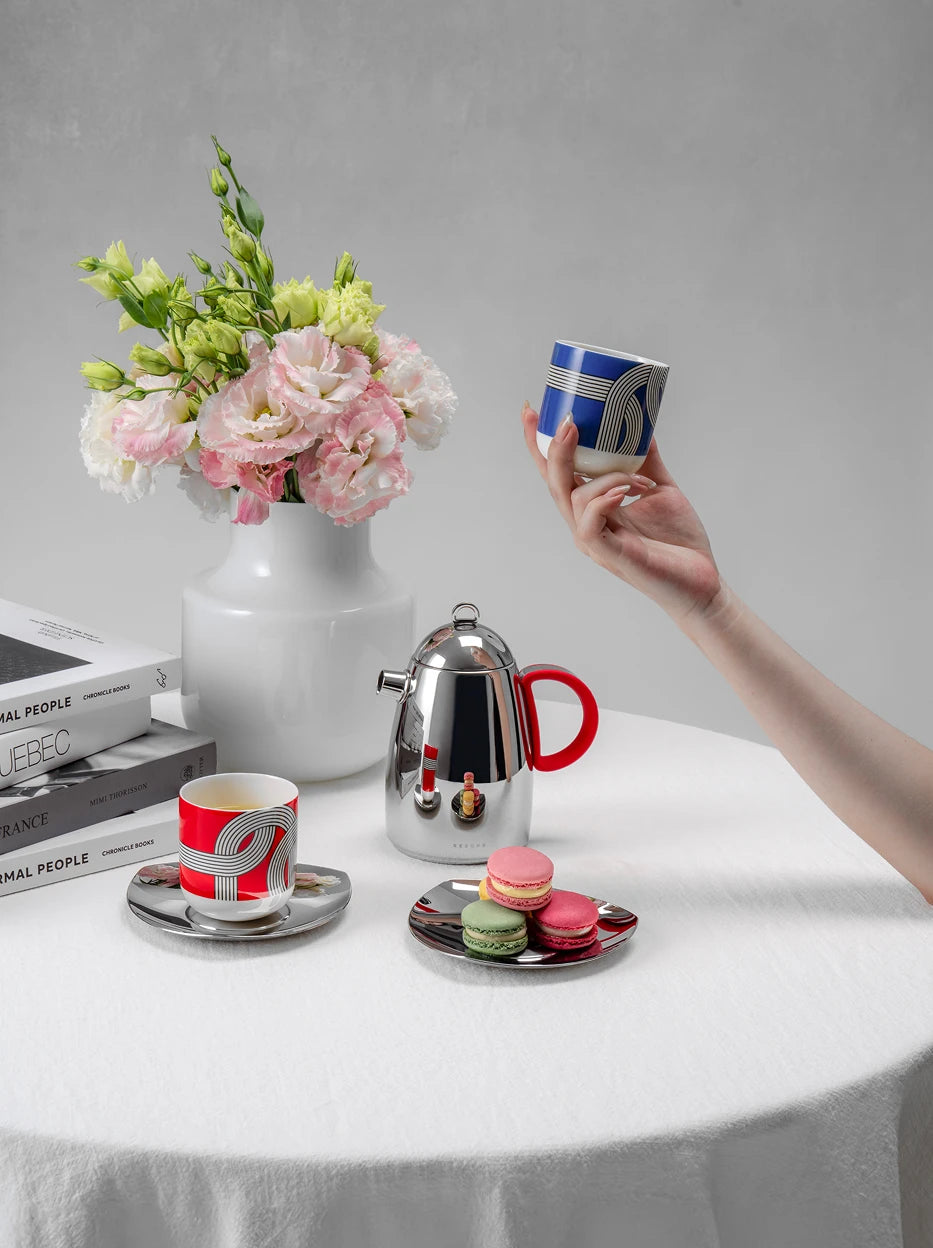 Cozy lifestyle shot of full ALK geometric print drinkware set on white round table, decorated with fresh carnation bouquet, stacked books and colorful macarons, light gray wall background
