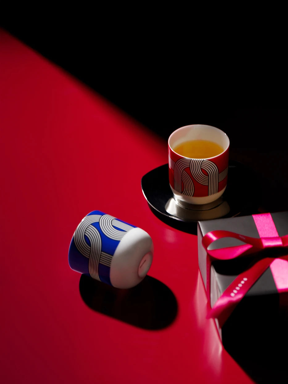 Luxury gift-themed still life of ALK red & blue geometric ceramic cups, glossy red-black split tabletop matched with ribbon gift box, one cup filled with golden tea liquid