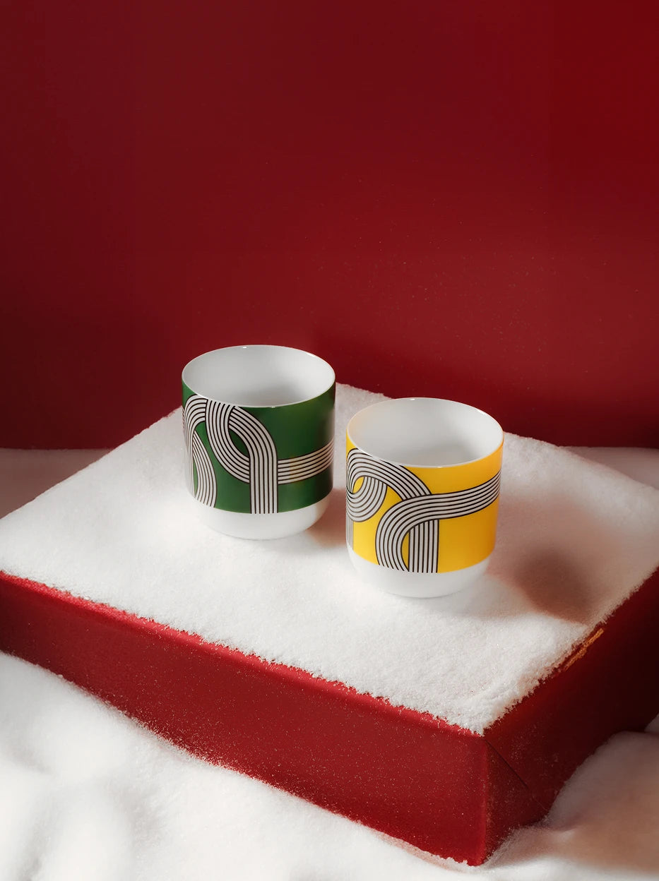 Festive winter lifestyle shot of ALK yellow and green geometric ceramic cups placed on red gift box covered with artificial snow, solid deep red background for holiday gift display