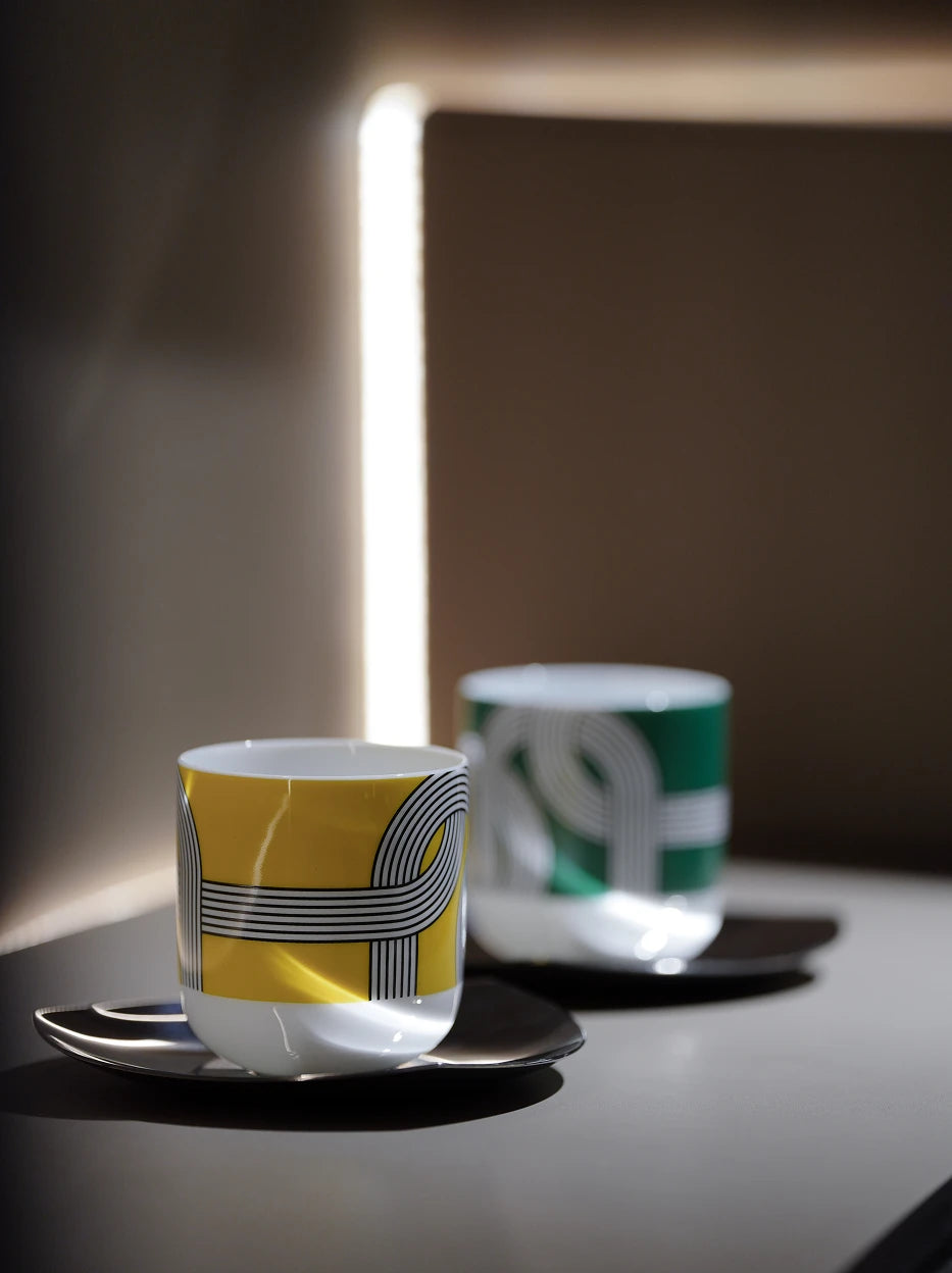 Minimal light-filled studio shot of ALK yellow & green ceramic cup set on light neutral side table, vertical soft light strip backdrop creating layered shadow texture