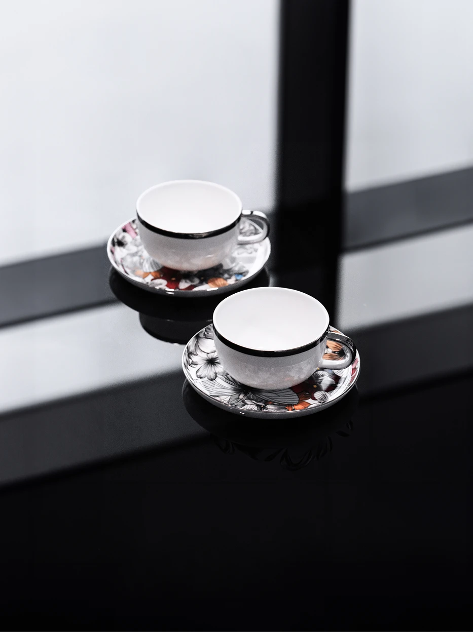 Minimalist studio display of dual BUTTERFLY WHISPER floral tea cup sets on reflective black glass table, frosted glass window background