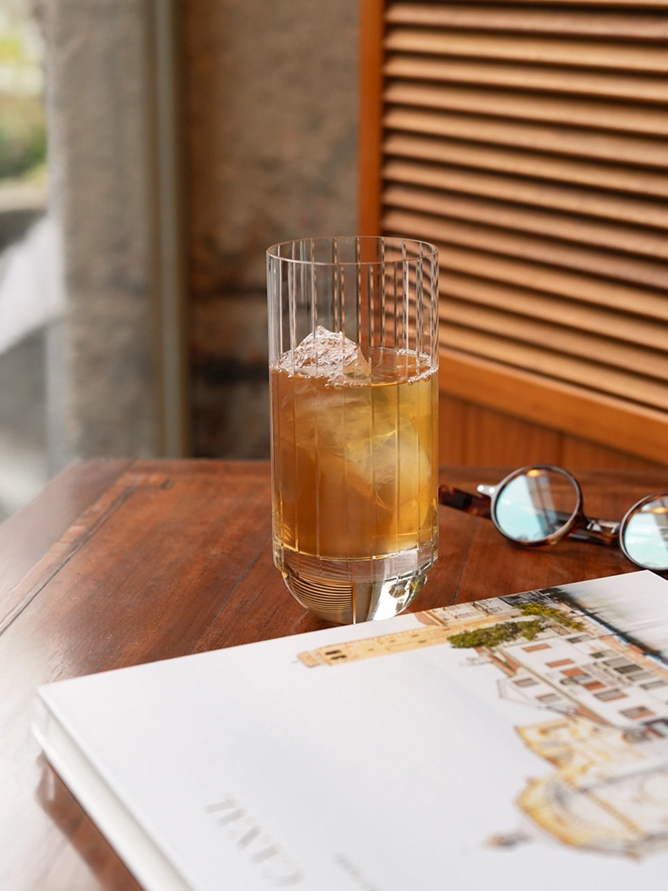 Lifestyle photo of RESONG HOME DEWDROPS Tall Glass, vertical ribbed tall crystal glass filled with iced cocktail on wooden table, minimalist home table decor