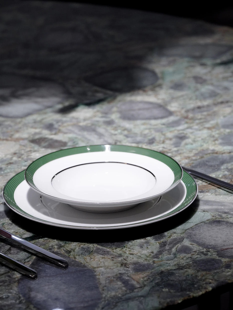 Minimal dark studio still life of stacked ERYA green rim deep plate set with silver cutlery on dark green marble tabletop