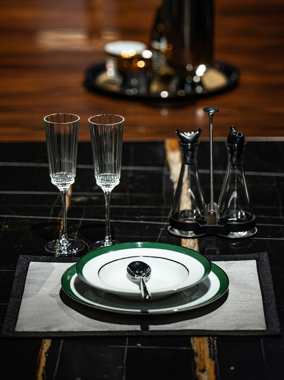Business dining table shot of ERYA green rim plate set with metal spoon, vertical striped champagne flutes and glass seasoning bottles on black wood textured tabletop